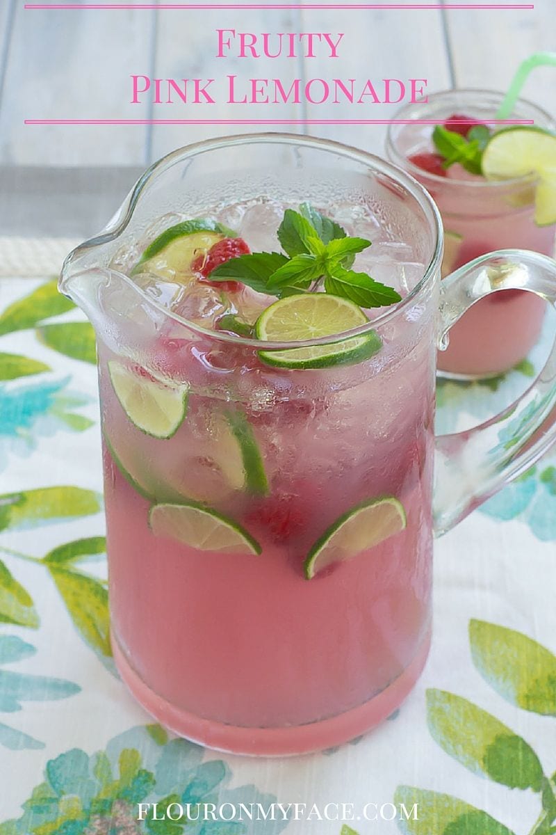 A glass pitcher filled with pink lemonade with berries, lime slices and ice cubes floating.