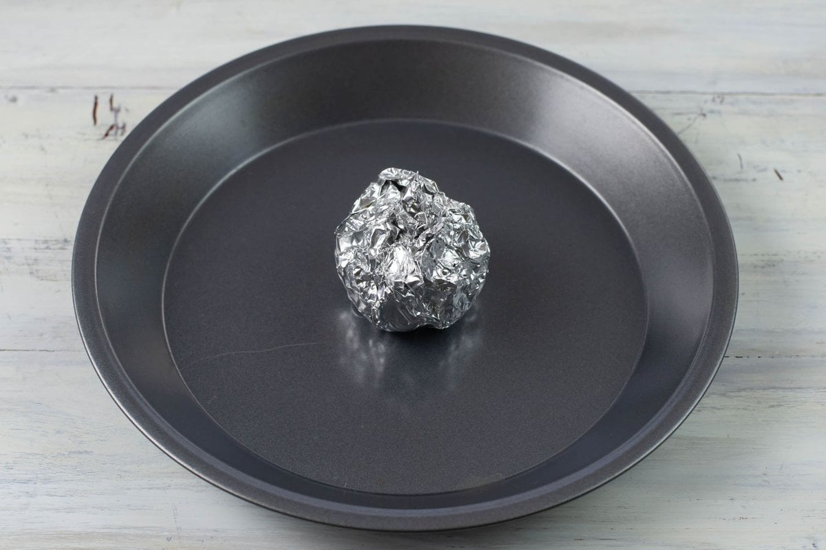 A whole head of garlic wrapped in aluminum foil in a metal pie plate.