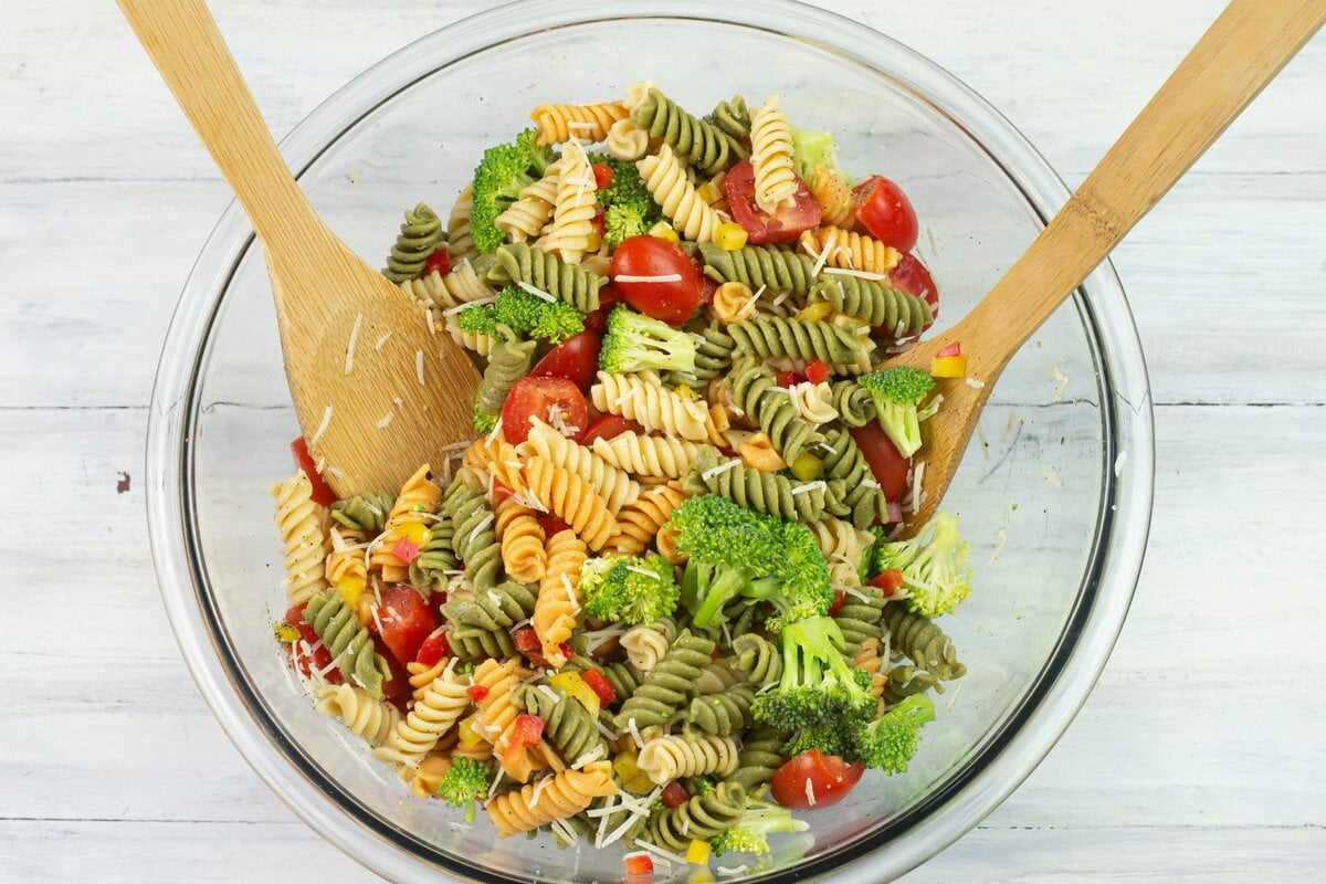 A bowl with cold pasta salad ingredients tossed together with wooden spoons.