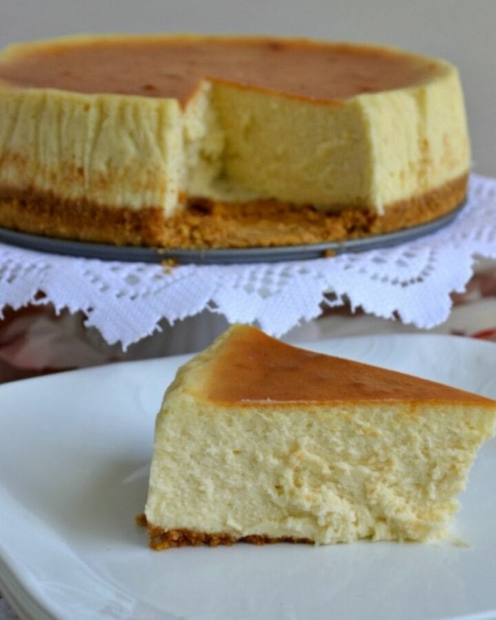 A slice of Vanilla Bean Cheesecake on a plate with the cheesecake in the background.