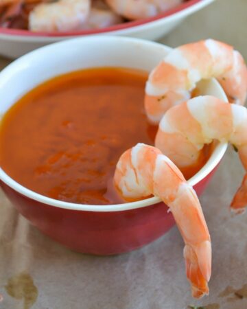 A small boil filled with spicy garlic dipping sauce with three boiled shrimp hanging over the edge.