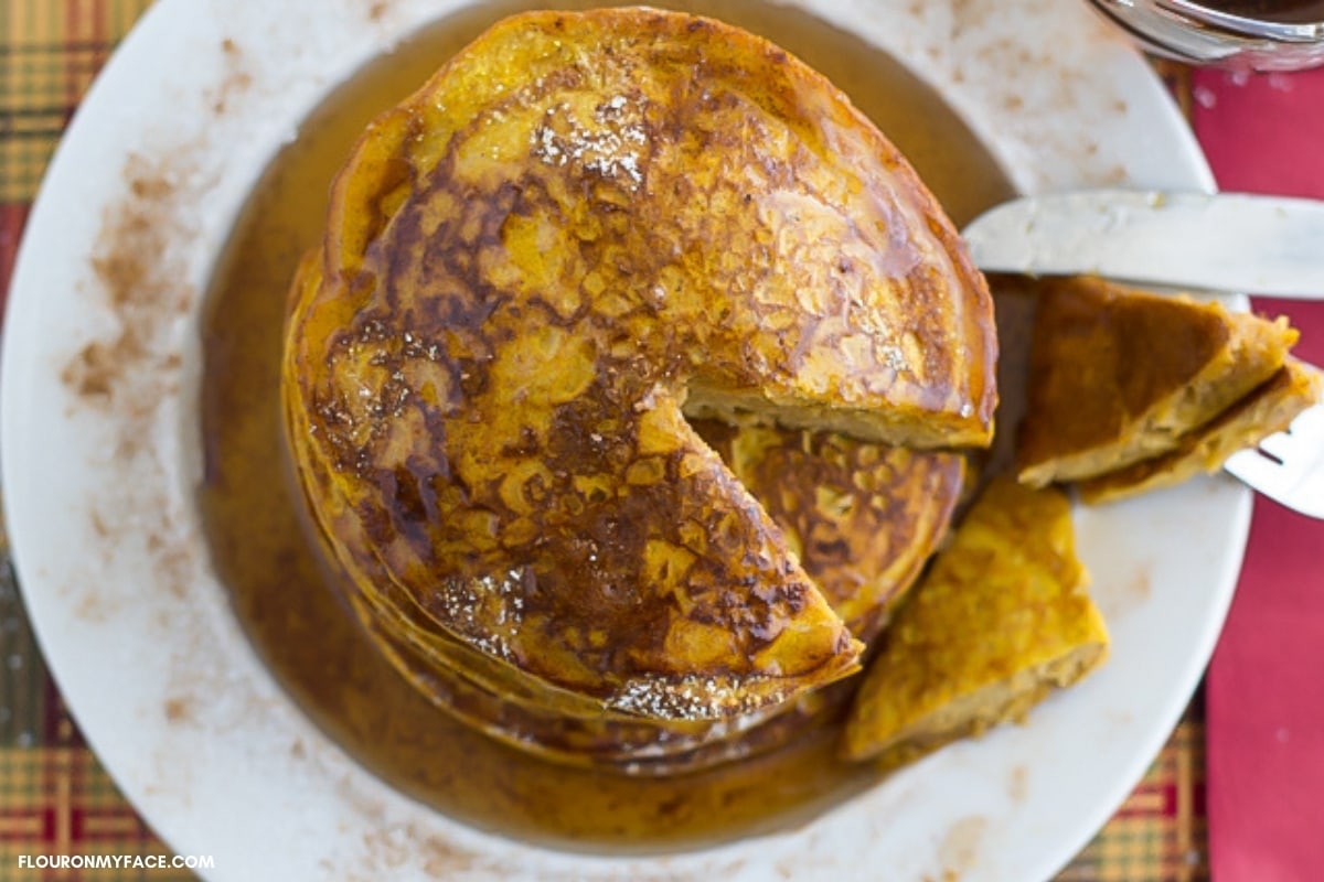 A stack of Pumpkin Spice Pancakes with syrup drizzled over the top and a piece cut. 