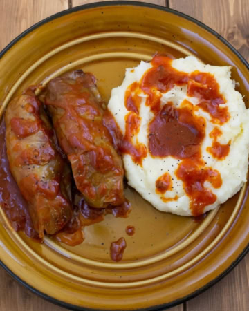 Stuffed cabbage rolls with a tomato based gravy served with mashed potatoes on a dinner plate.