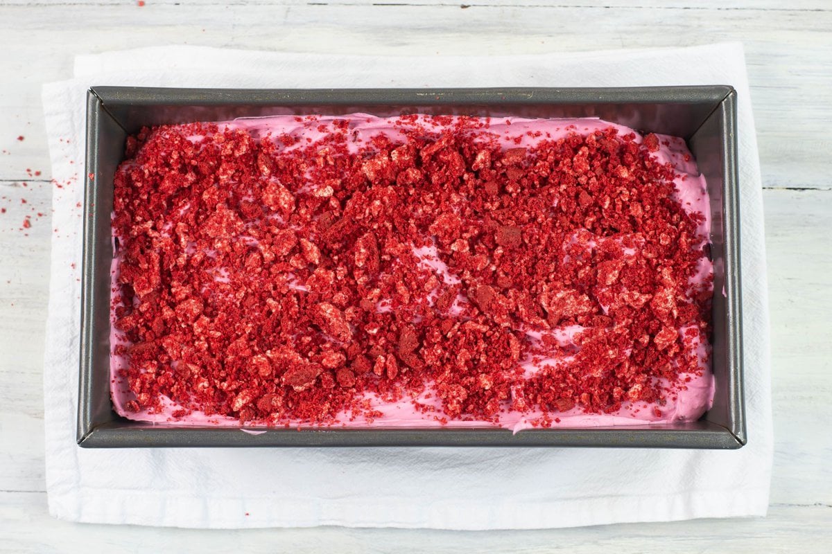 Crushed red velvet cookies sprinkled over no churn ice cream in a pan before freezing.