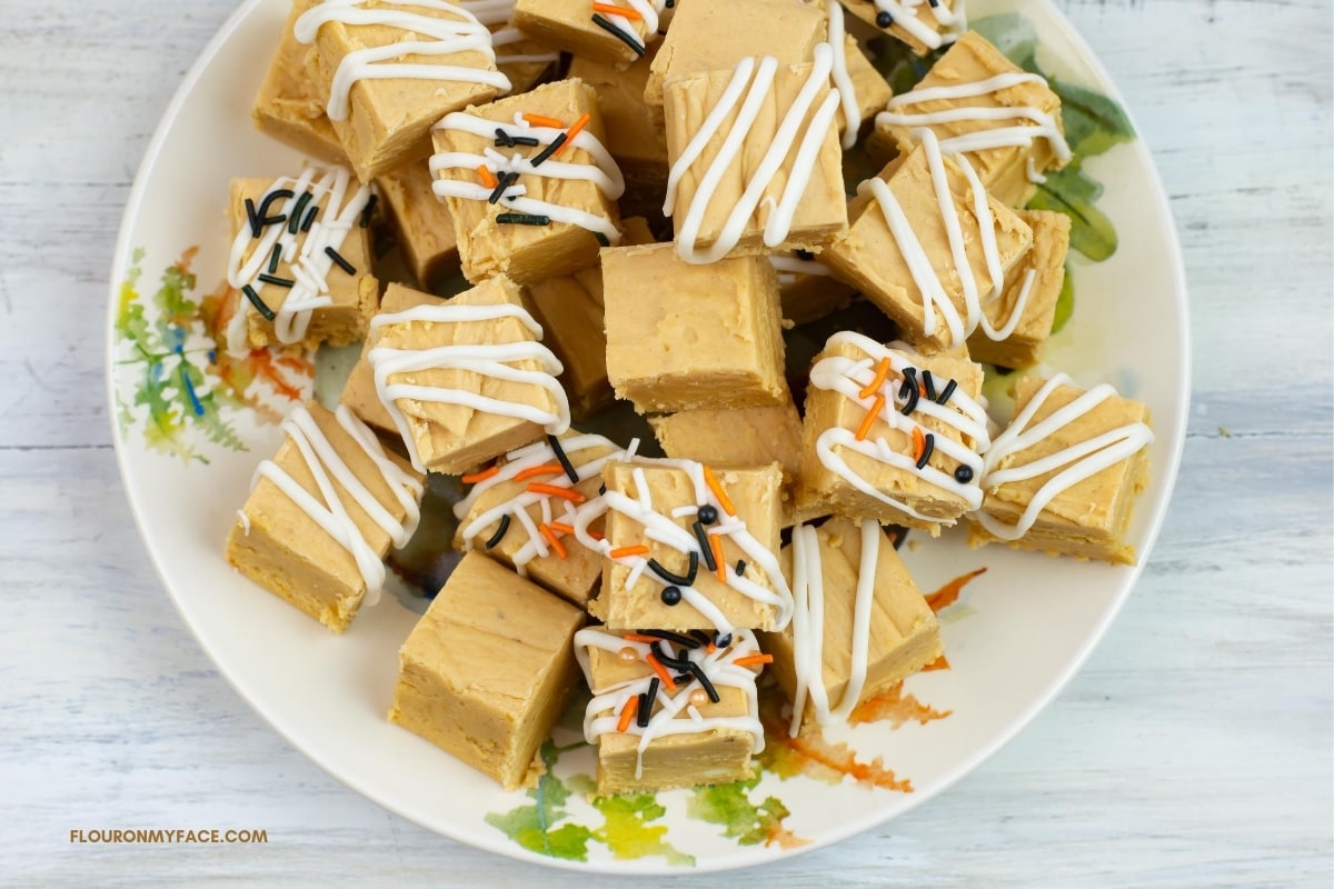 a fall dessert plate with a pile of pumpkin fudge cut into squares decorated with drizzled white choclate and orange and black sprinkles