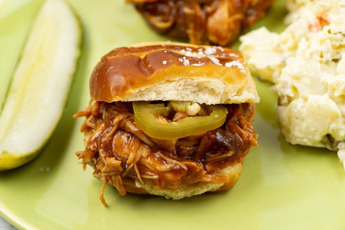 Bitesize pulled chicken sandwich served with a pickle and potato salad on a green dinner plate.
