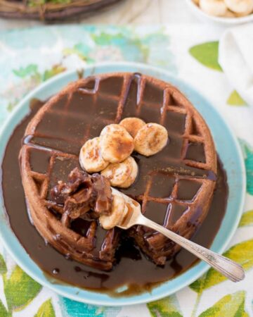 A stack of Nutella Waffles topped with sliced banana and drizzles with syrup.