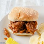 Sweet and Tangy Crock Pot Pulled Pork Sandwich recipe served with chips, pickles, and homemade coleslaw