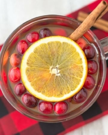 Hot Apple Cider Christmas Wassail recipe served in a mug on a black and red plaid cloth napkin with fresh cranberries, an orange slice and a cinnamon stick for garnish.