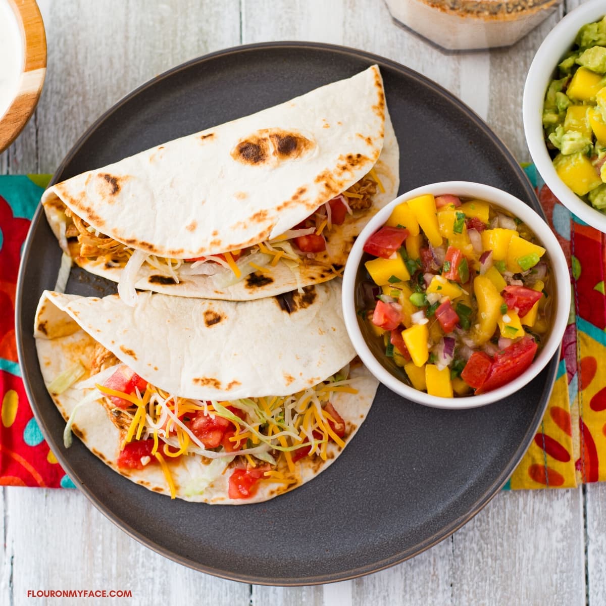 a grey dinner plate with 2 chicken tacos made with flour tortillas topped with shredded cheese, lettuce, and homemade mango salsa