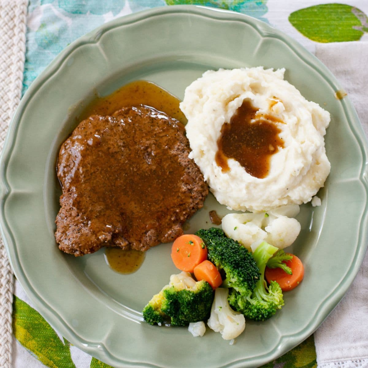 A serving of Cube Steak smothered in gravy with mashed potatoes and vegetables on a dinner plate.
