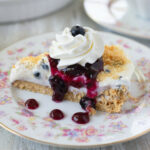 sliced no bake blueberry cheesecake pie on a floral patterned dessert dish with whipped cream and blueberry sauce drizzled over it.