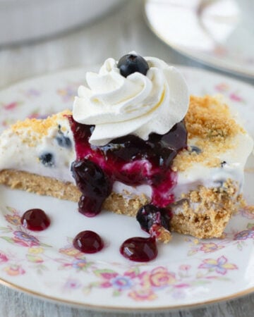 sliced no bake blueberry cheesecake pie on a floral patterned dessert dish with whipped cream and blueberry sauce drizzled over it.
