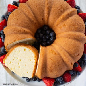A Pound Cake with a slice cut out served on a cake plate with fresh strawberries and blueberries encircling the cake.