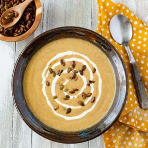 a brown pottery bowl filled with creamy pumpkin soup garnished with maple pumpkin seeds and a swirl of sour cream