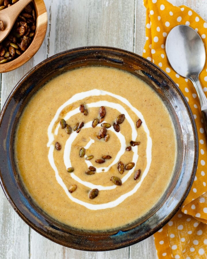 a brown pottery bowl filled with creamy pumpkin soup garnished with maple pumpkin seeds and a swirl of sour cream