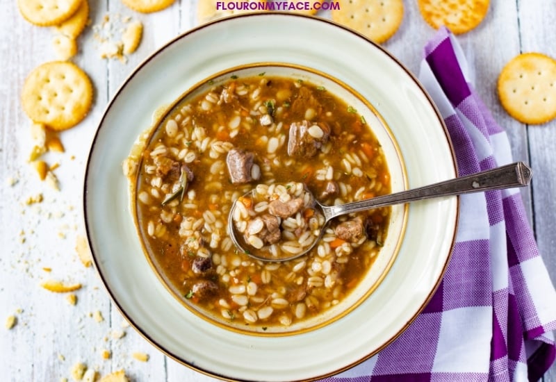 A bowl of Instant Pot Beef Barley Soup with a purple and white cloth napkin and Ritz crackers