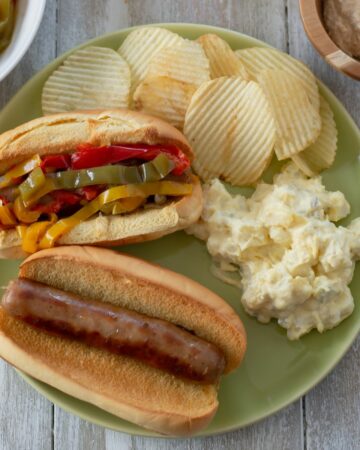 Brats on a bun with peppers served with chips and potato salad.