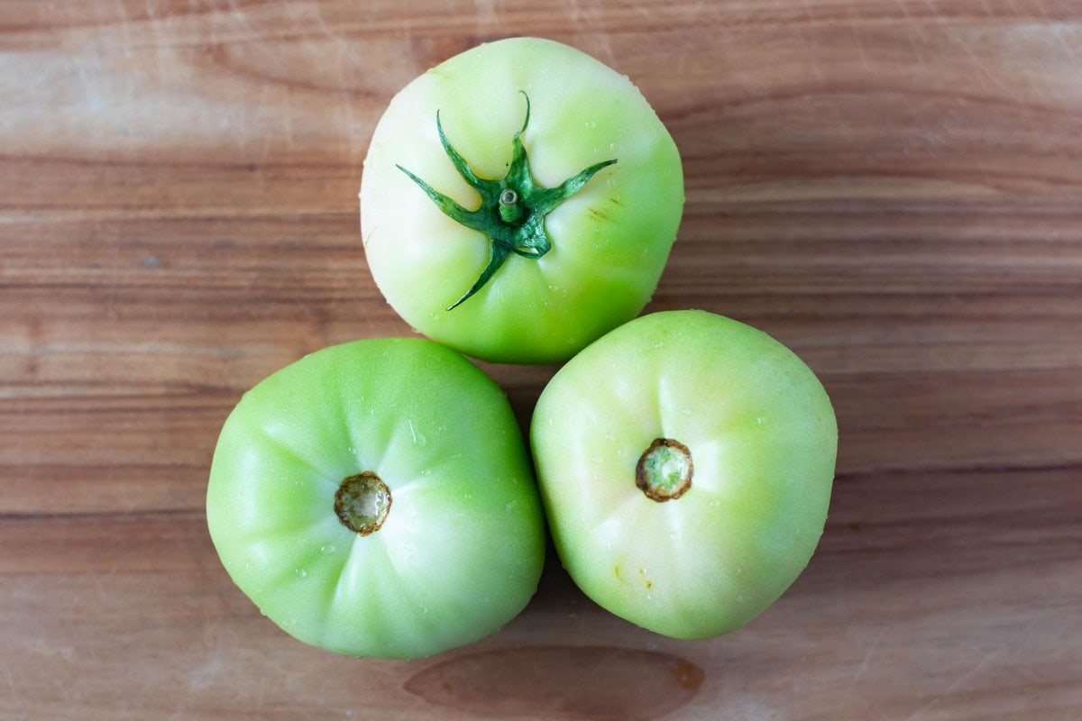 Three large green tomatoes on wooden cutting board.