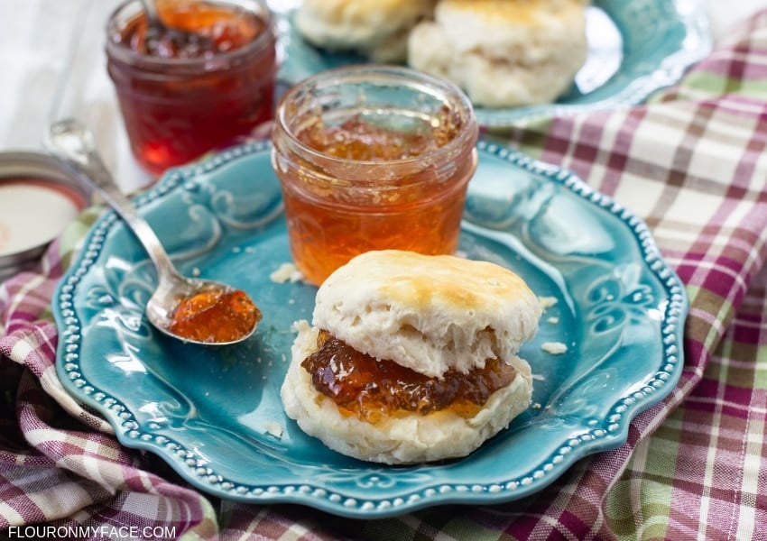A plate with a fresh biscuit with a thick spread of homemade Scuppernong Jelly with a jar of the jelly on a cloth napkin.