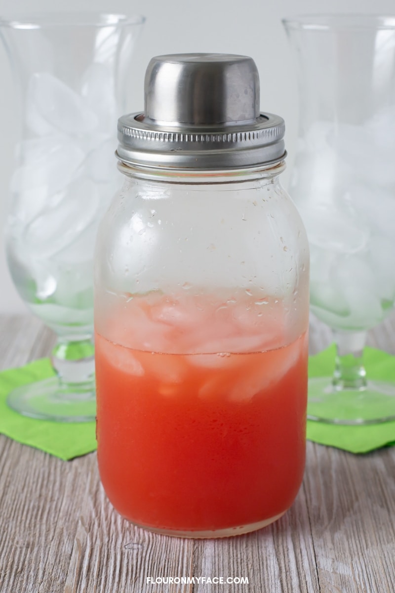 Close up photo of a clear Mason jar cocktail shaker filled with a classic Hurricane with two hurricane cocktail glasses in the background.