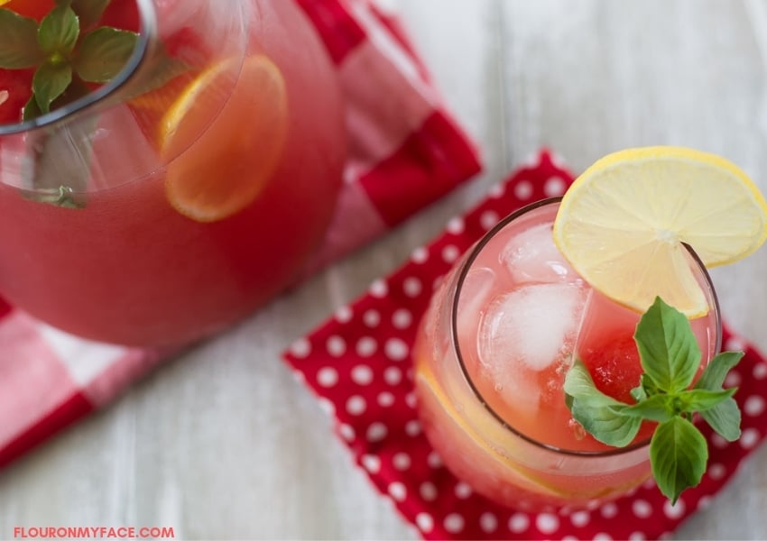 a glass and a pitcher filled with watermelon lemonade with a lemon twist and a sprig of fresh basil