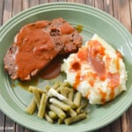 A green plate with 2 slices of Crock Pot Meatloaf covered in a tomato soup gravy with a side of mashed potatoes and green beans.