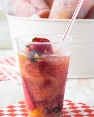 front view photo of a single Frozen Fruit Punch Cups on a checked nakin with a straw. In the background is a white metal bucket filled with ice with 6 individual Frozen Fruit Cups for serving