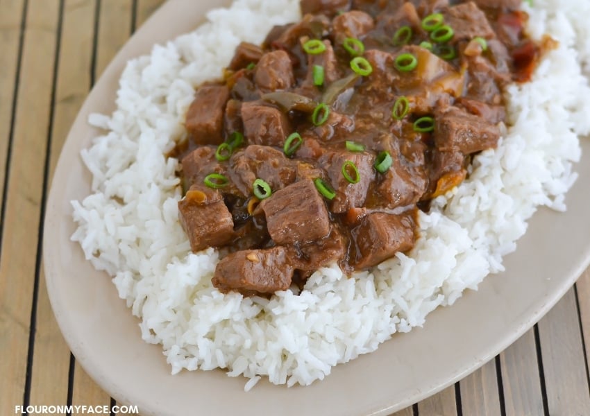  Crock Pot Pepper Steak served over a bed of cooked white rice on an oval platter with green onions sprinkled on top.