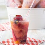 front view photo of a single Frozen Fruit Punch Cups on a checked nakin with a straw. In the background is a white metal bucket filled with ice with 6 individual Frozen Fruit Cups for serving