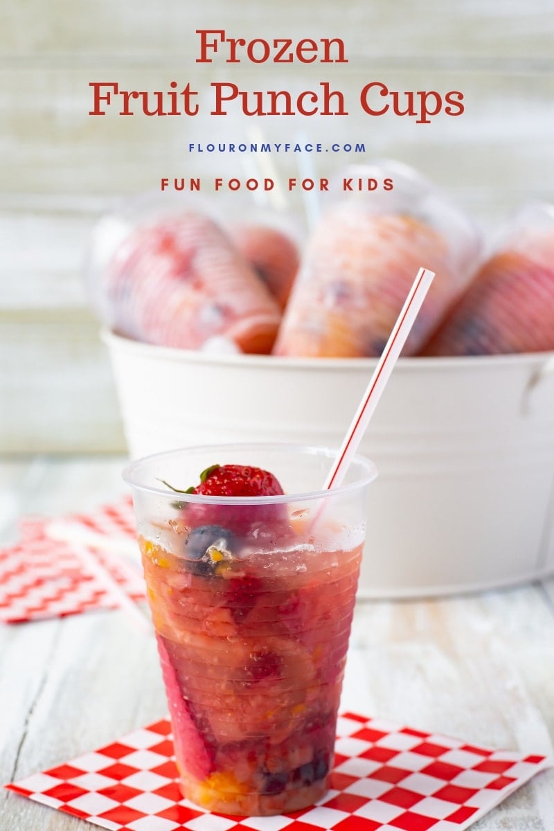 A clear plastic cup filled with fruit and punch then frozen. With a metal bucket in the background filled with ice and more Frozen Fruit Punch Cups chilling for a summer frozen treat.