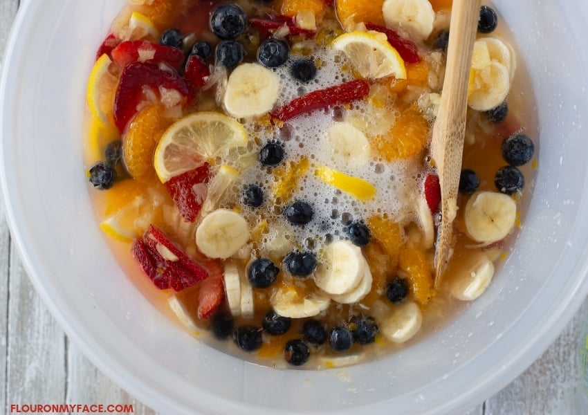 A plastic bowl filled with sliced bananas, fresh blueberries, mandarin orange slices, sliced strawberries, crushed pineapple, sugar, and lemon lime soda for a Frozen Fruit Punch Cups recipe.