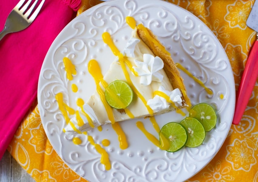Above the plate photo of a slice of Mango Key Lime Pie that has been garnished with whipped cream, a slice of lime and a drizzle of mango sauce on top of a yellow cloth napkin.