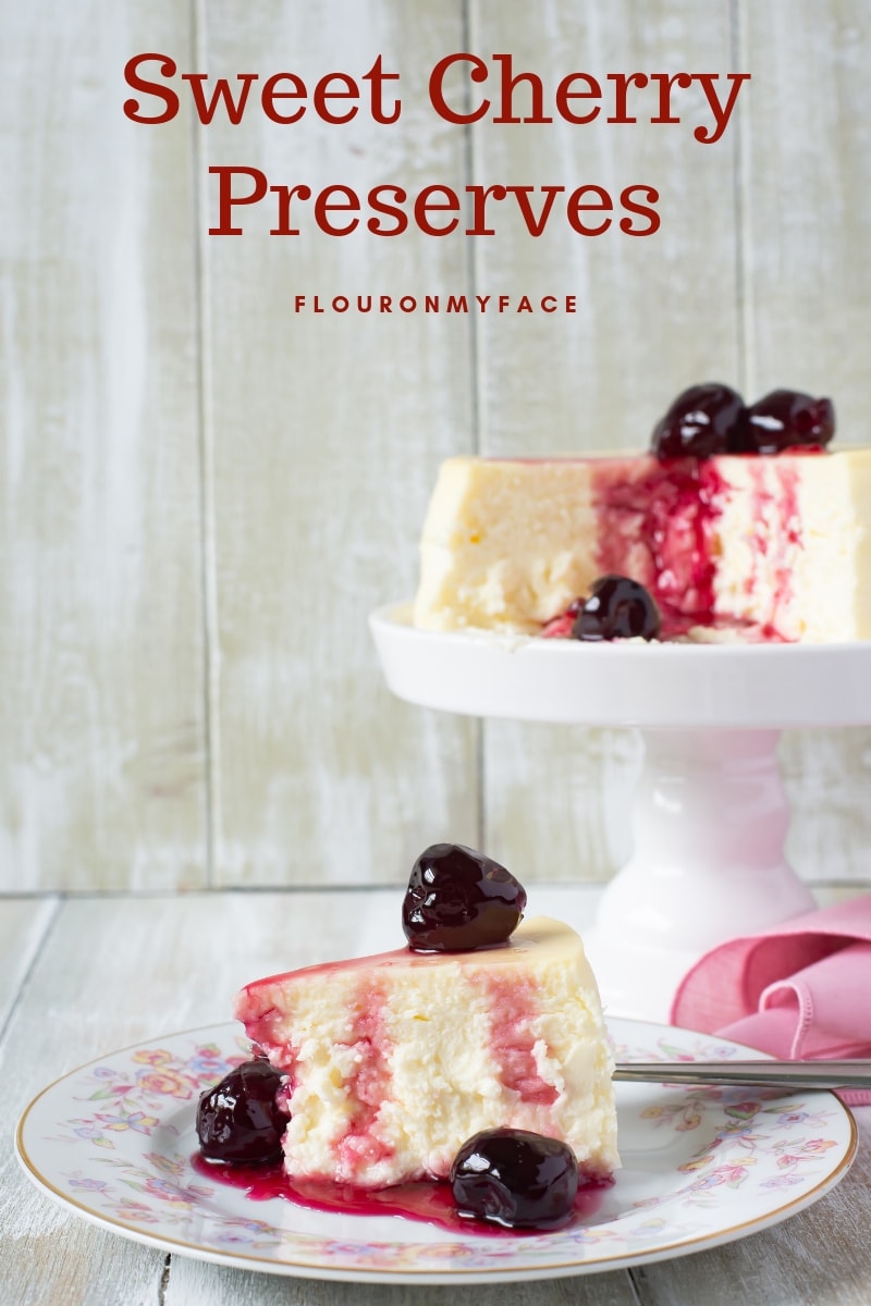 A New York cheese cake topped with homemade Cherry Preserves on a cake stand , with a dessert plate with one slice of cheesecake with a spoonful of homemade cherry preserves drizzled over the top.