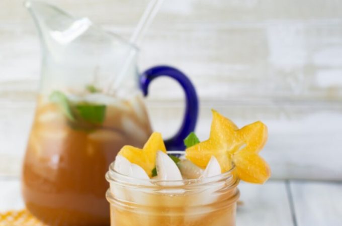 A mason jar glass filled with Carambola iced tea recipe with sliced starfruit and mint garnish. The filled glass pitcher is in the background.