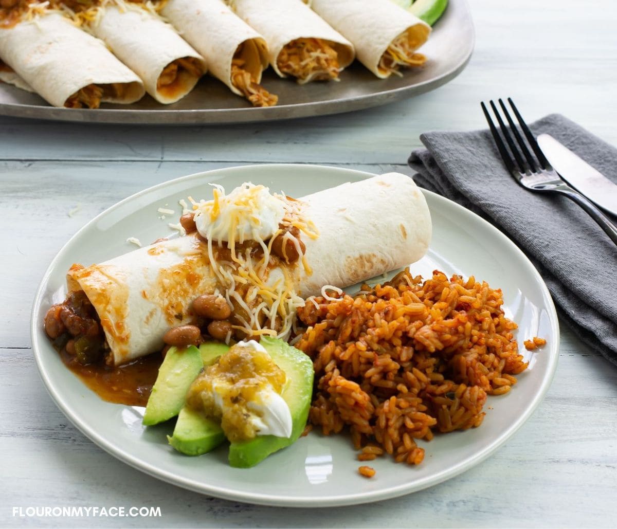 A dinner plate with chicken burritos, slice avocado and rice.