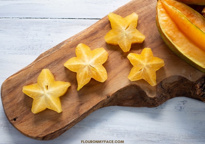 A wooden cutting board with 4 pieces of sliced star fruit with a whole fruit in the background.