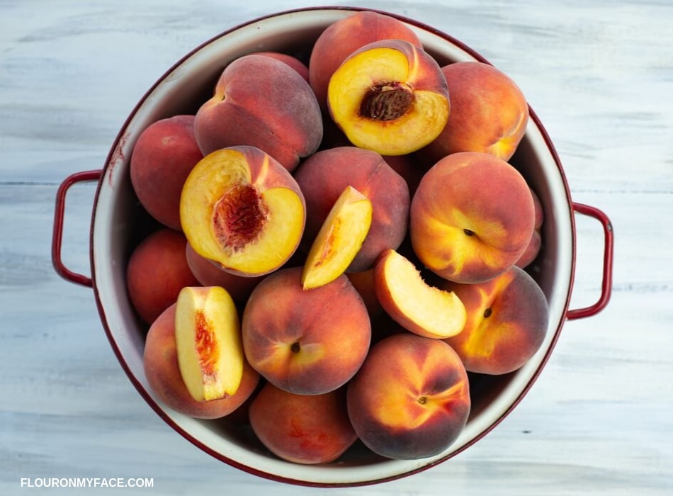 Fresh summer peaches piled high in a white enamel colander, with a few pieces of a sliced peach on top of the whole peaches.