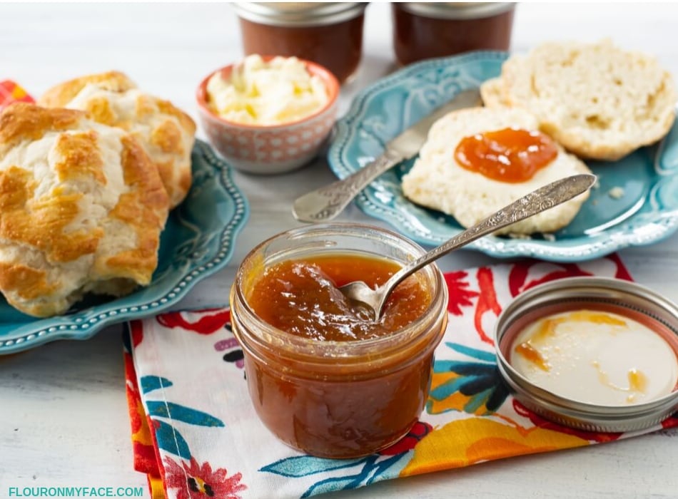 A table setting with biscuits on a plate in the background with a opened jar of thick and rich homemade Peach Honey Butter.