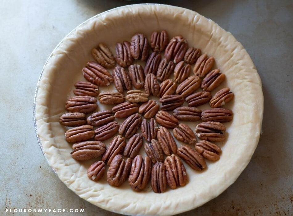 an overhead photo of an unbaked pie crust with a layer of pecan halve arranged in a pretty pattern.