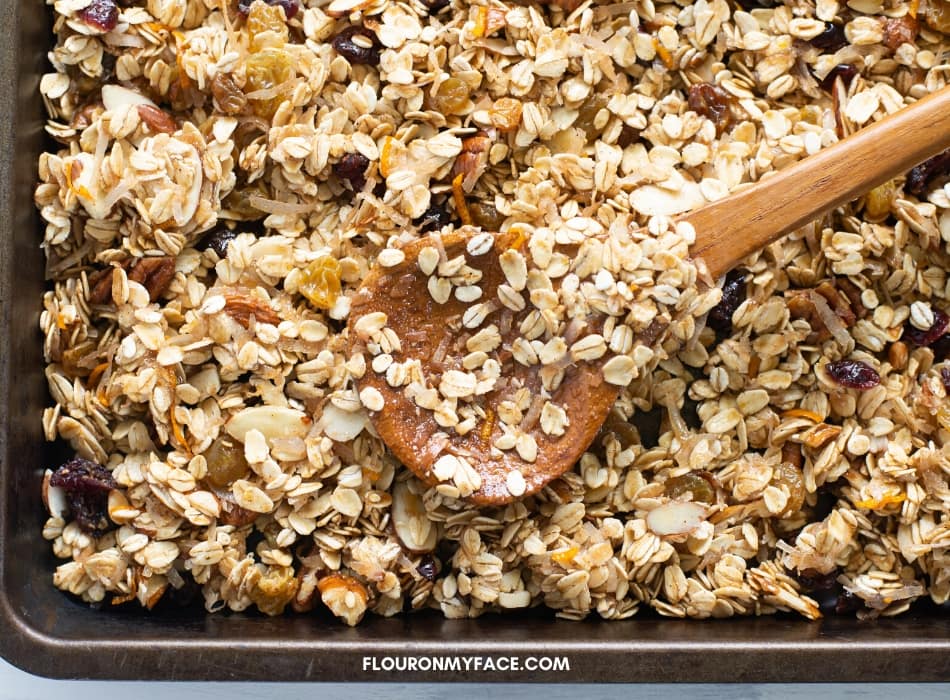 unbaked Christmas Granola spread with a wooden spoon over a large baking sheet before it is browned in the oven