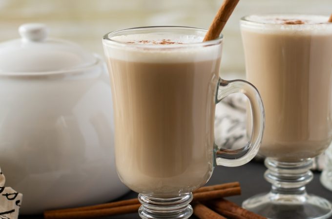 A mug of Crock Pot Chai Latte topped with frothy milk. cinnamon and garnished with a cinnamon stick. A white tea pot is in the background