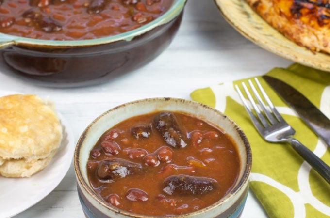 A serving bowl of Crock Pot Cowboy Beans in a small serving bowl on a table with a plate of grilled chicken, corn and a biscuit.