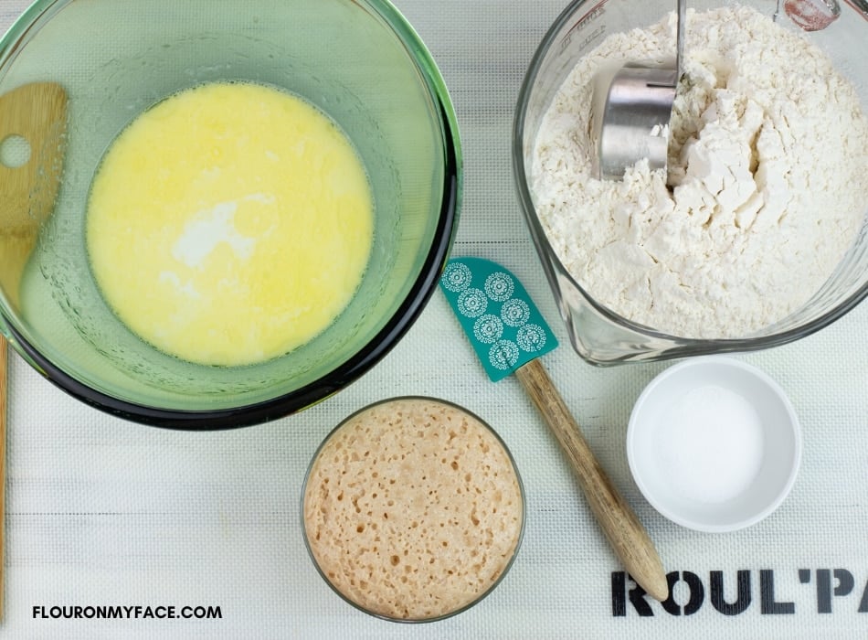 Homemade bread ingredients on a work surface: a bowl with mild and melted butter, a bowl of flour and salt, a small bowl of proofed yeast, 