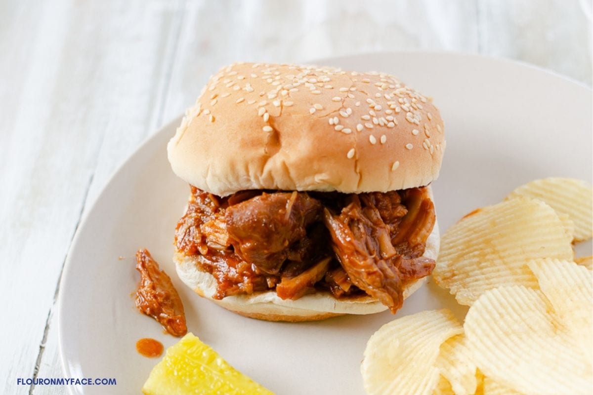 Pulled pork sandwich on a plate with chips and a pickle.