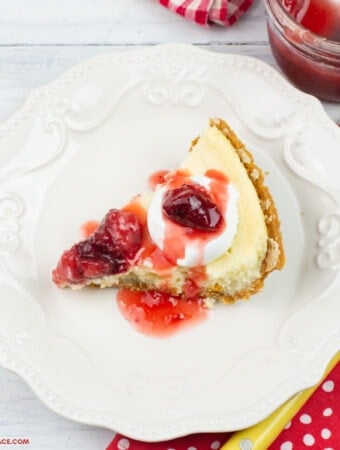 A slice of homemade cheesecake topped with homemade strawberry topping on a light beige dessert plate with a red and white polka dot napkin, a fork, a mason jar with strawberry topping in the background