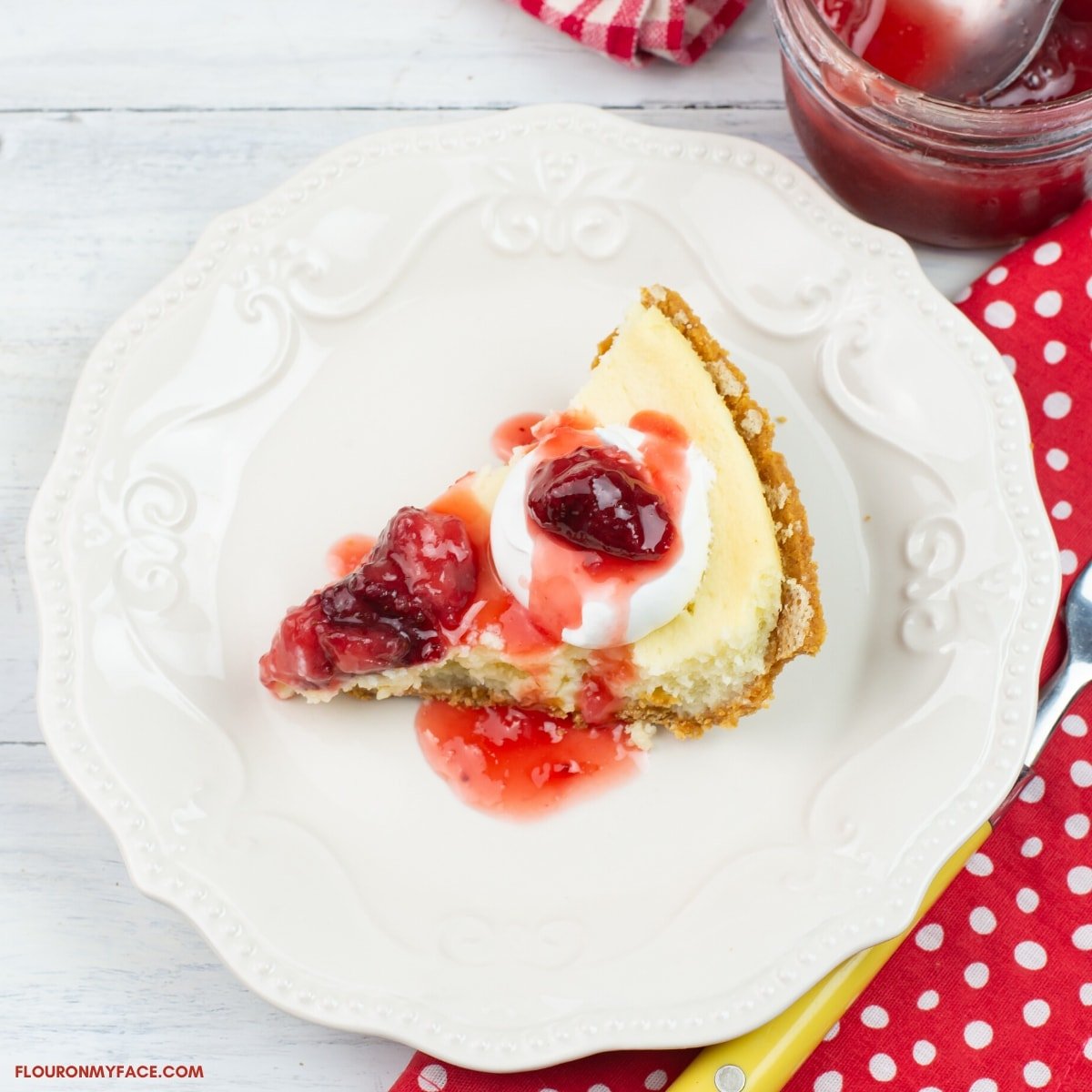 A slice of homemade cheesecake topped with homemade strawberry topping on a light beige dessert plate with a red and white polka dot napkin, a fork, a mason jar with strawberry topping in the background