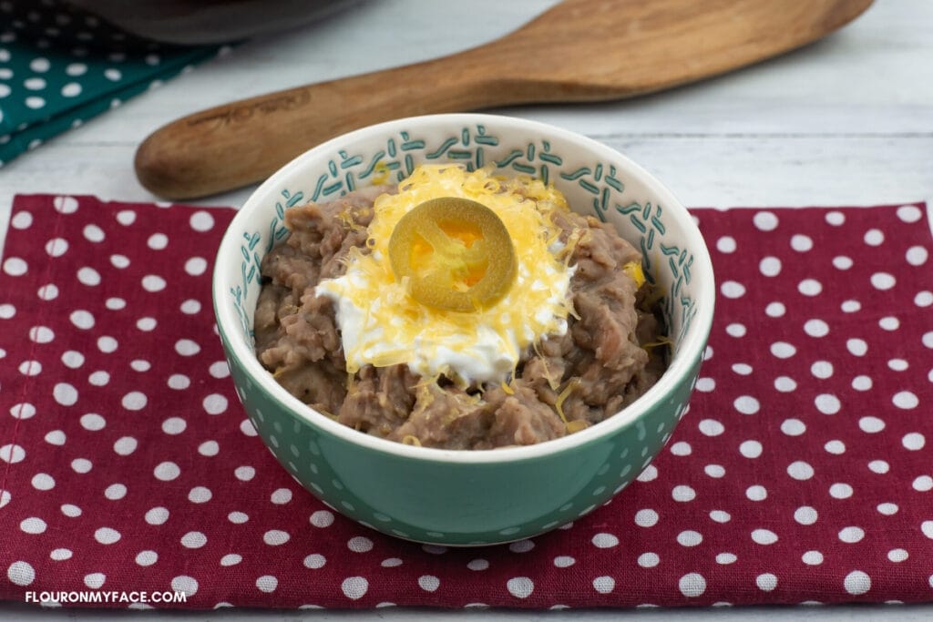 a small teal serving bowl filled with homemade refried beans made in the Instant Pot topped with a dollop of sour cream, shredded cheese and a slice of jalapeno pepper