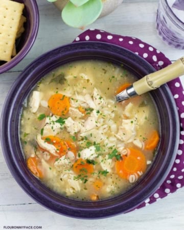 Pressure cooked chicken and barely soup in a purple fiesta ware bowl with a bowl of crackers and a glass of water