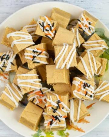 A dessert plate with squares of white chocolate pumpkin fudge stacked in a pile. Some of the fudge has been decorated with drizzled white chocolate and orange, black, and white sprinkles for fall.
